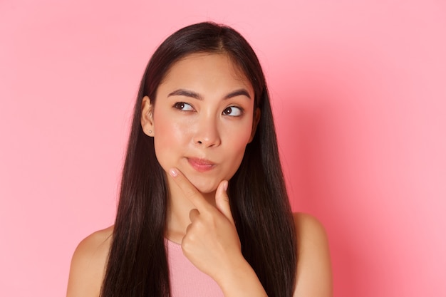 Beauty, fashion and lifestyle concept. Close-up of thoughtful asian trendy girl, making plan, choosing something, touching chin and looking upper left corner while thinking, pink wall