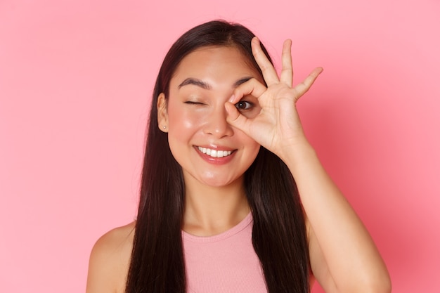 Beauty, fashion and lifestyle concept. portrait of kawaii attractive asian girl showing okay gesture over eye and winking carefree, smiling pleased, guarantee quality, recommend place.