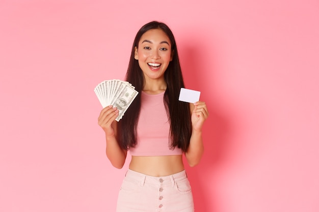 Beauty, fashion and lifestyle concept. Waist-up of young beautiful asian woman in trendy outfit showing credit card and money, smiling as explain how to make purchase, pink wall.