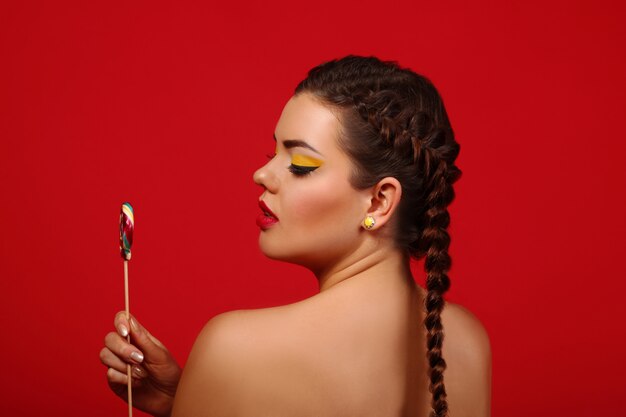 Beauty fashion model girl eating colorful lollipop. Surprised funny young woman with braids hairstyle, nails and makeup beige, isolated on a red wall