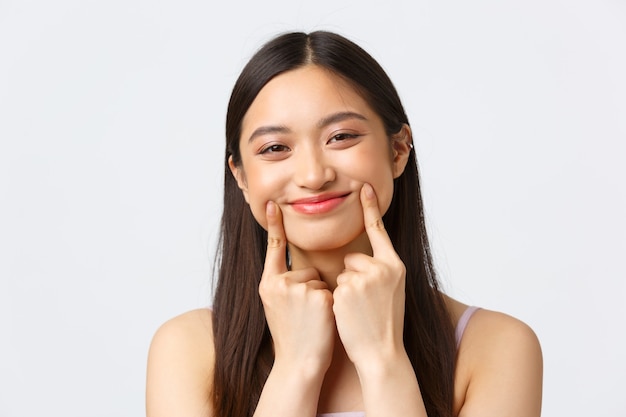 Beauty, fashion and people emotions concept. Close-up portrait of adorable silly asian girl in dress, pulling happy smile with fingers over lips