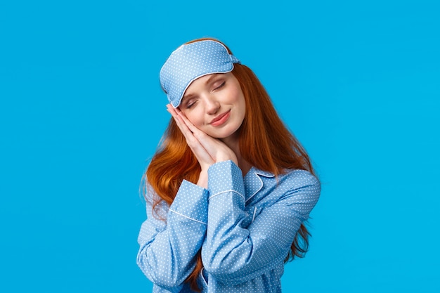 Beauty, female and lifestyle concept. Relaxed and happy charming redhead woman in nightwear, sleep mask, having sweet dreams, lean on palms like pillow, smiling with closed eyes