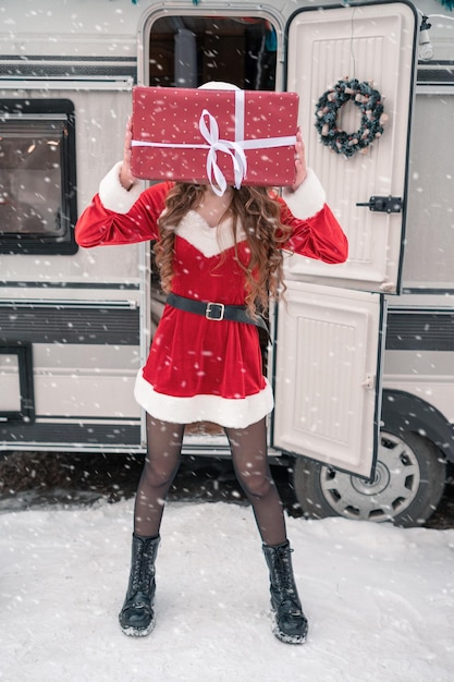 Красавица в костюме санты с подарочной коробкой в зимнем кемпинге готовится к новому году
