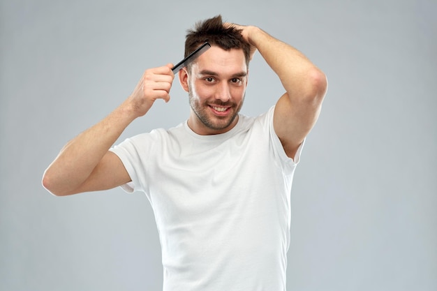 beauty, grooming and people concept - smiling young man brushing hair with comb over gray background