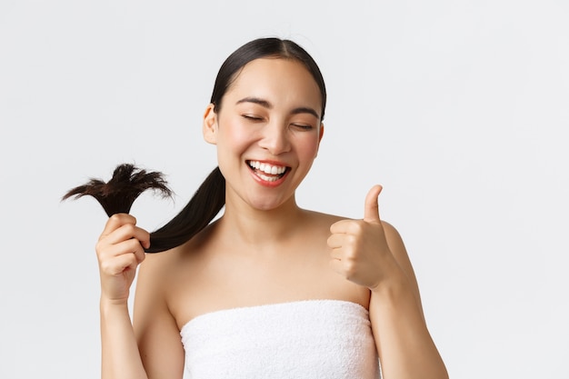 美容、脱毛製品、シャンプー、ヘアケアのコンセプトです。満足して幸せなアジアの女の子の親指のアップと健康な髪の端を示すバスタオルで満足している白い壁に立っているクローズアップ。