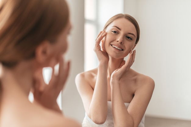 Beauty and her reflection in a mirror. Over the shoulder view of attractive woman touching her face and smiling while spending time at home