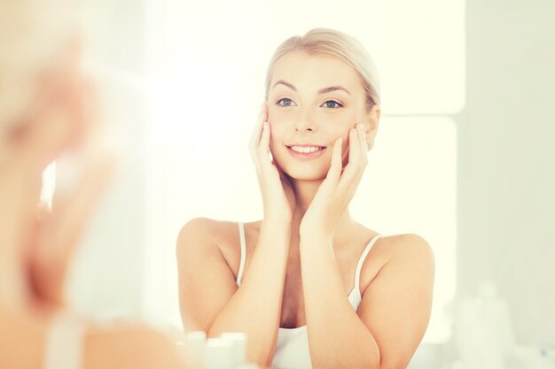 beauty, hygiene, morning and people concept - smiling young woman looking to mirror at home bathroom