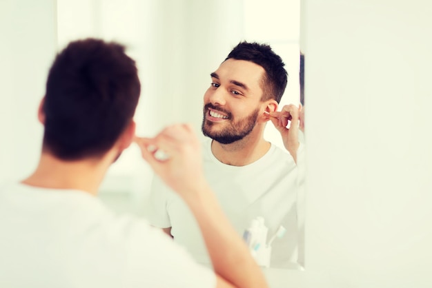 beauty, hygiene and people concept - smiling young man cleaning ear with cotton swab and looking to mirror at home bathroom