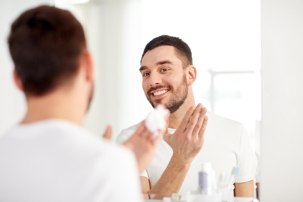 美しさ、衛生、シェービング、グルーミング、人々 のコンセプト - 自宅の浴室で鏡を見て、顔にシェービング フォームを適用する笑顔の若い男