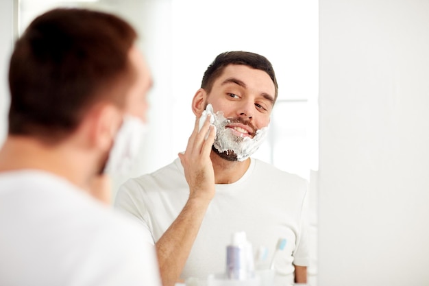 красота, гигиена, бритье, уход и концепция людей - улыбающийся молодой человек смотрит в зеркало и наносит пену для бритья на лицо в домашней ванной