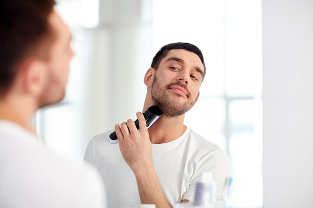 beauty, hygiene, shaving, grooming and people concept - young man looking to mirror and shaving beard with trimmer or electric shaver at home bathroom