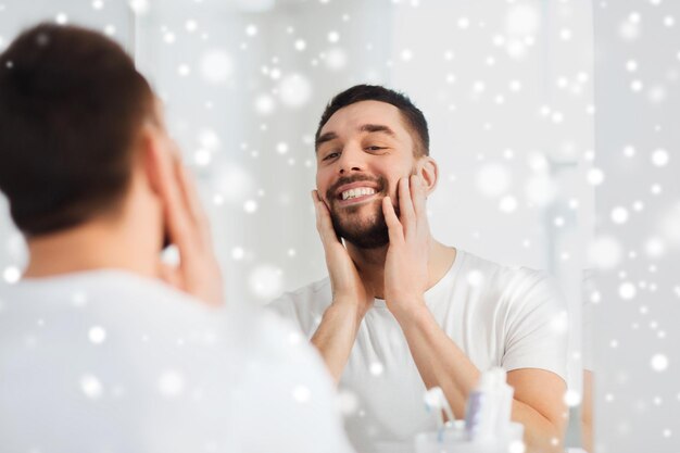 beauty, hygiene, shaving and people concept - smiling young man looking to mirror at home bathroom over snow