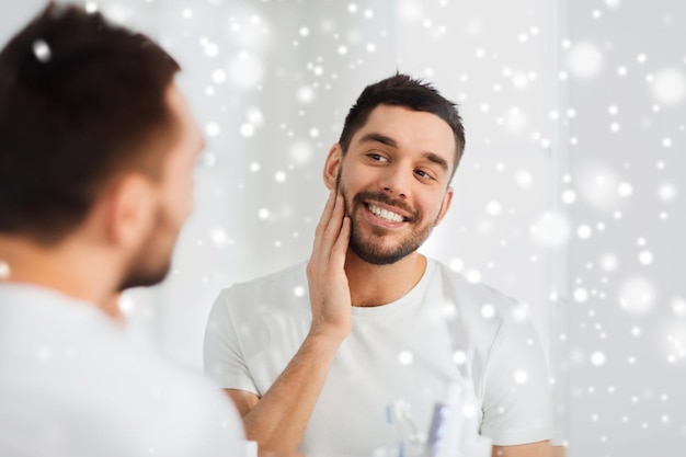 美しさ、衛生、髭剃り、冬、人のコンセプト – 雪の上で自宅のバスルームの鏡を見ている笑顔の若い男性
