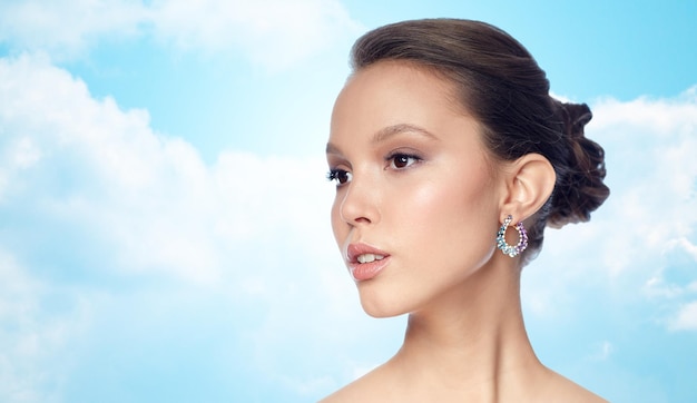 Foto concetto di bellezza, gioielli, accessori, persone e lusso - primo piano del volto di una bella donna asiatica con orecchini su sfondo blu cielo e nuvole