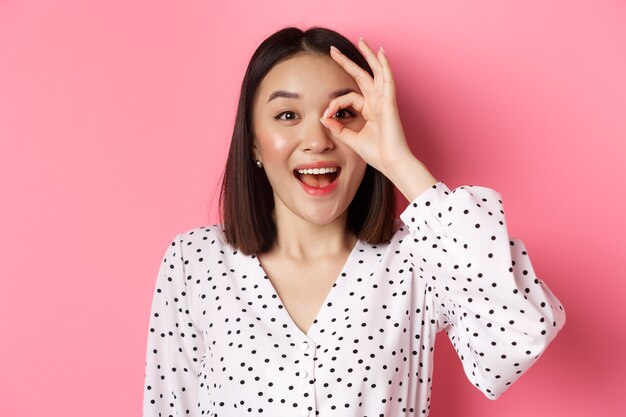 Beauty and lifestyle concept. Beautiful asian girl looking through okay sign and smiling amazed, standing against pink background.