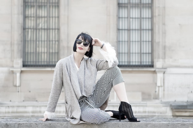 Beauty look style Woman sit on stone fence on house facade Fashion vogue concept Girl with brunette hair red lips in sunglasses Model in high heel shoes fashionable clothes parisian woman