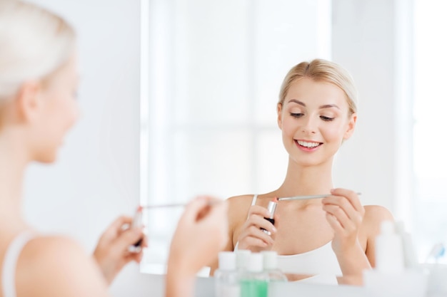 Foto concetto di bellezza, trucco, cosmetici, mattina e persone - giovane donna sorridente con rossetto e applicatore che applica il trucco e guarda allo specchio nel bagno di casa