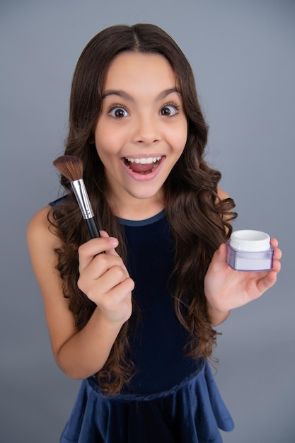 Beauty make up and kids cosmetics Teenage girl with brush applying blush to face isolated on grey studio background Excited face cheerful emotions of teenager girl