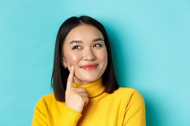 Beauty and makeup concept. close up of pensive asian girl smiling, imaging something and looking at upper left corner, pitching an idea, standing over blue background.