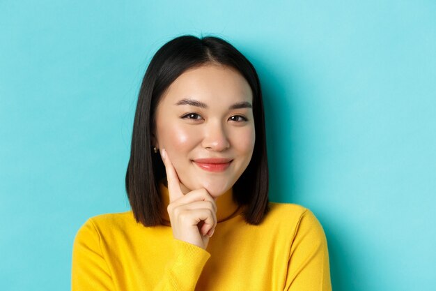 Beauty and makeup concept. Close up of thoughtful asian woman looking pleased at camera and smile, having an idea, standing over blue background.