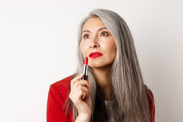 Concetto di bellezza e trucco. sensuale femmina asiatica matura con i capelli grigi, guardando da parte e mostrando rossetto rosso, in piedi su sfondo bianco.