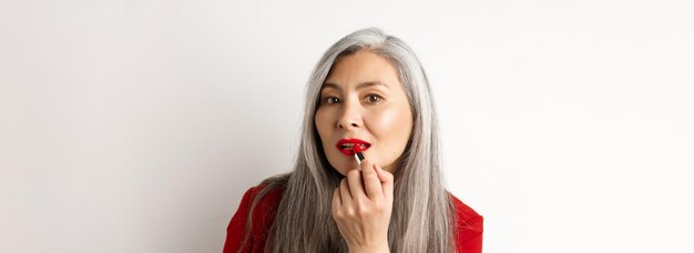 美しさと化粧のコンセプト スタイリッシュなアジアの成熟した女性の灰色の髪を鏡で見て赤を適用します。