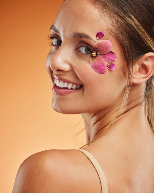 Beauty makeup and creative with woman petals on face for design flowers and natural cosmetics against orange background in studio Spring skincare and smile with girl for color model and orchid