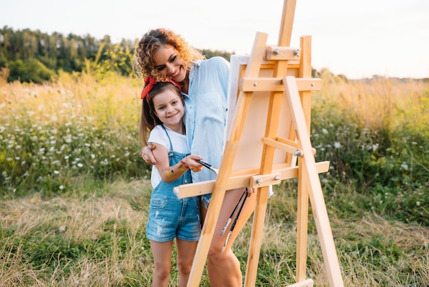 Pittura della madre di bellezza con la sua piccola figlia. donna alla moda che disegna l'immagine con la bambina. ragazzo carino in una maglietta bianca e jeans blu.