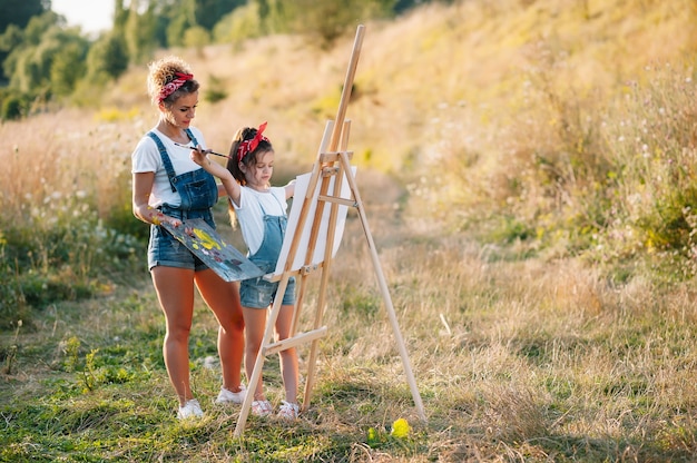 Pittura della madre di bellezza con la sua piccola figlia. donna alla moda che disegna l'immagine con la bambina. ragazzo carino in una maglietta bianca e jeans blu.