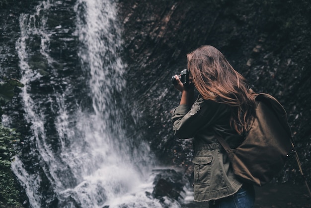 자연의 아름다움을 포착해야 합니다. 배낭을 메고 폭포 근처에 서서 물을 촬영하는 젊은 현대 여성