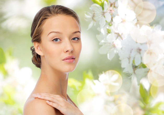 Beauty, people, body care and health concept - smiling young woman face and hand on bare shoulder over green natural cherry blossom background