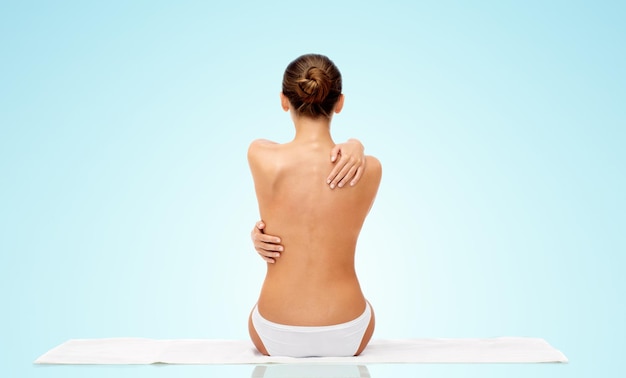 beauty, people and bodycare concept - beautiful topless young woman in white panties sitting on towel from back over blue background