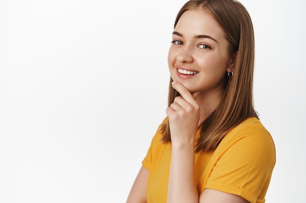 Beauty and people concept. Close up portrait of attractive young woman smiling pleased, touch chin and look thoughtful at camera, standing confident against white background