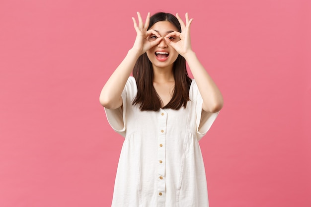 Beauty, people emotions and summer leisure concept. Playful gorgeous funny asian girl in white dress, make fake glasses from hands and looking through it at super cool promo, smiling amazed