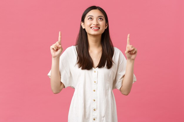 Bellezza, emozioni delle persone e tempo libero estivo e concetto di vacanza. sognante donna asiatica tenera e felice in abito bianco, che guarda e punta le dita con un sorriso compiaciuto leggendo il banner promozionale.