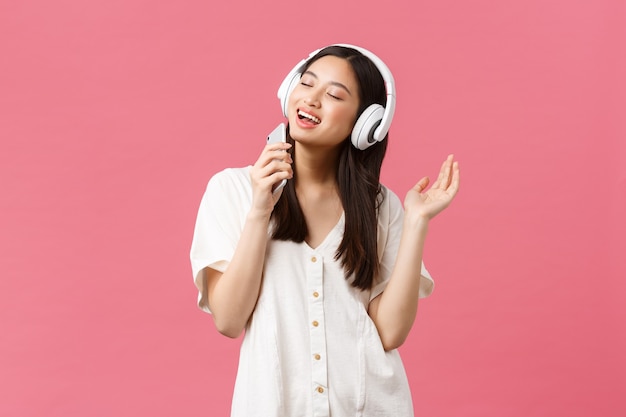 Bellezza, emozioni delle persone e concetto di tecnologia. ragazza asiatica felice e spensierata che utilizza l'applicazione karaoke del telefono cellulare, canta nel microfono dello smartphone, ascolta musica in cuffia, sfondo rosa