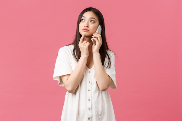 Beauty, people emotions and technology concept. Thoughtful asian girl making order delivery using smartphone, thinking while talking over phone, looking up, pondering as choosing, pink background