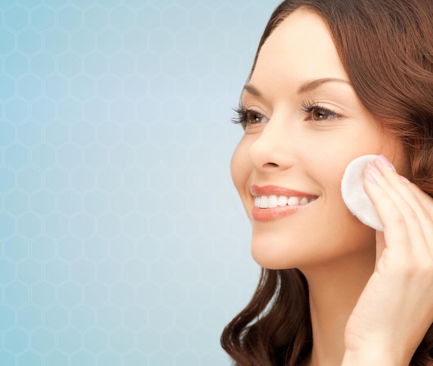 beauty, people and health concept - beautiful smiling woman cleaning face skin with cotton pad over blue background