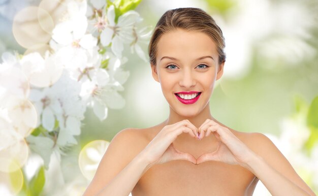 Photo beauty, people, love, valentines day and make up concept - smiling young woman with pink lipstick on lips showing heart shape hand sign over green natural background with cherry blossom