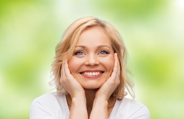 beauty, people and skincare concept - smiling middle aged woman in white shirt touching face over green natural background