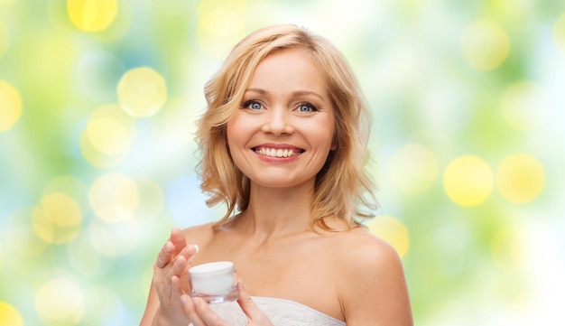 Photo beauty, people, skincare and cosmetics concept - happy middle aged woman with cream jar over green holidays lights background