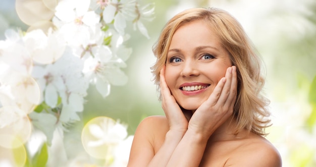 beauty, people, skincare and natural cosmetics concept - smiling woman with bare shoulders touching face over cherry blossom background