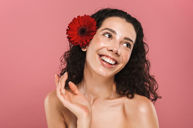 Beauty photo of nude happy woman 20s with long hair and no makeup holding red flower, isolated over pink wall