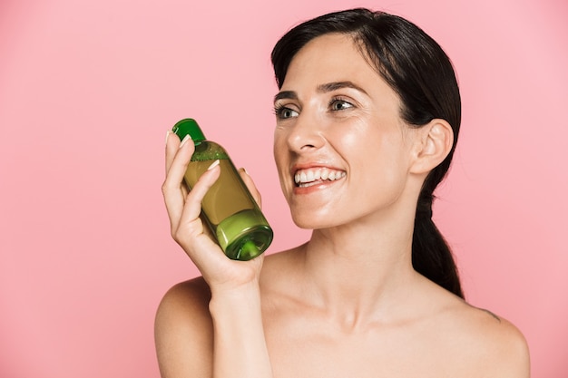 Beauty portrait of an attractive happy healthy topless brunette woman isolated over pink wall, showing cosmetic bottle