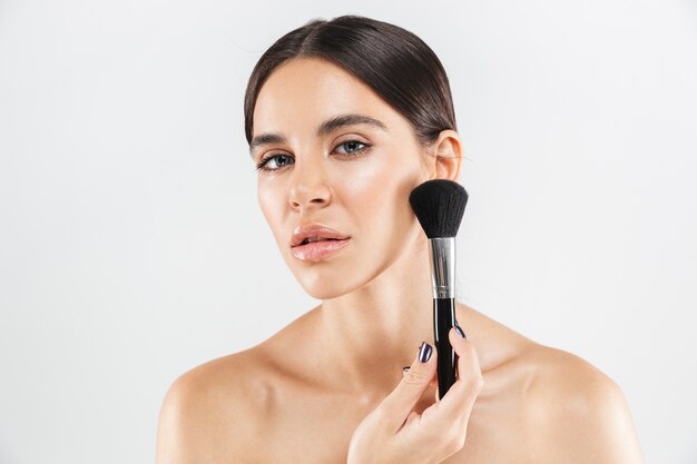 Beauty portrait of an attractive healthy woman standing isolated over white wall, applying makeup with a brush