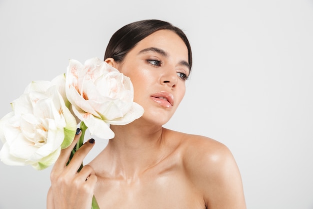 Beauty portrait of an attractive sensual healthy woman standing isolated over white wall, posing with a flower