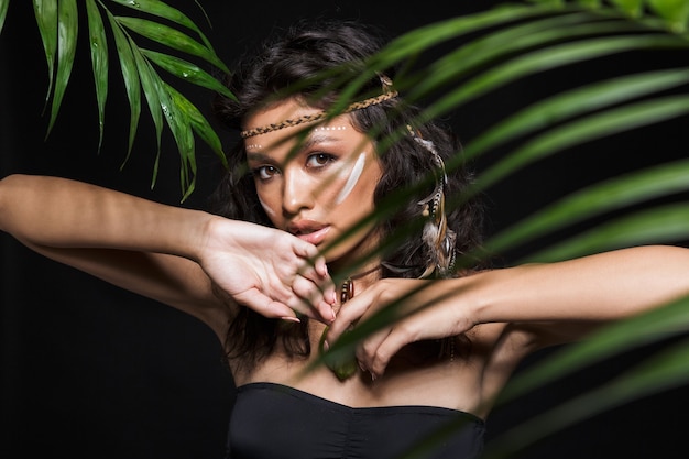 Beauty portrait of an attractive sensual young brunette woman wearing tribal accessories posing with tropical leaves isolated