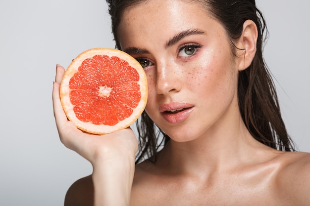 Beauty portrait of an attractive sensual young woman with wet brunette long hair standing isolated on gray, showing halved grapefruit, posing