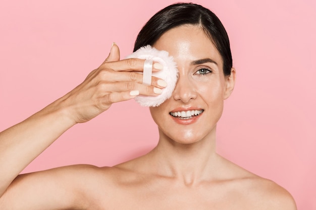 Beauty portrait of an attractive smiling young topless woman standing isolated over pink wall, using powder puff