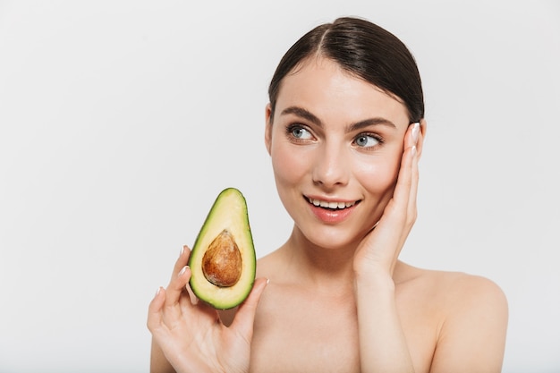 Beauty portrait of an attractive young brunette woman standing isolated over white wall, showing sliced avocado